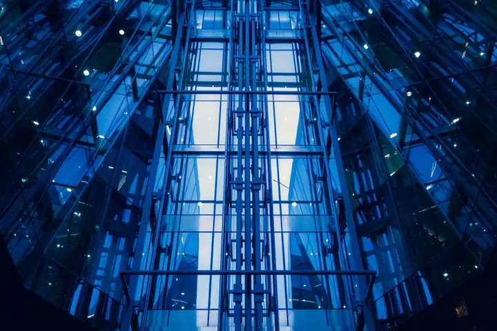 Poço de elevadores panorâmicos em vidro e estrutura metálica azul, visto de baixo