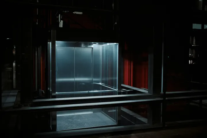 Cabina de elevador iluminada, em vidro e metal, num ambiente industrial escuro